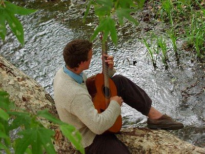 A man sitting down at a river playing a guitar.